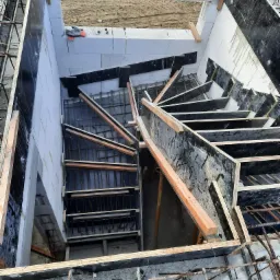 Budowa schod&oacute;w spiralnych w domu murowanym, widok z g&oacute;ry na szalunek z drewnianymi wzmocnieniami i zbrojenie stalowe, bloczki z betonu kom&oacute;rkowego w tle.
