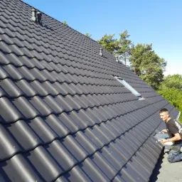 Dekarz na kolanach montuje ciemnoszarą dachówkę na skośnym dachu z oknem dachowym, widoczne kominki wentylacyjne i drzewa w tle pod błękitnym niebem.