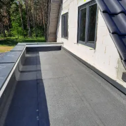 Widok z góry na płaski dach pokryty papą bitumiczną, przylegający do ściany budynku z oknami i fragmentem dachu spadzistego z czarną dachówką, w tle widoczny las.