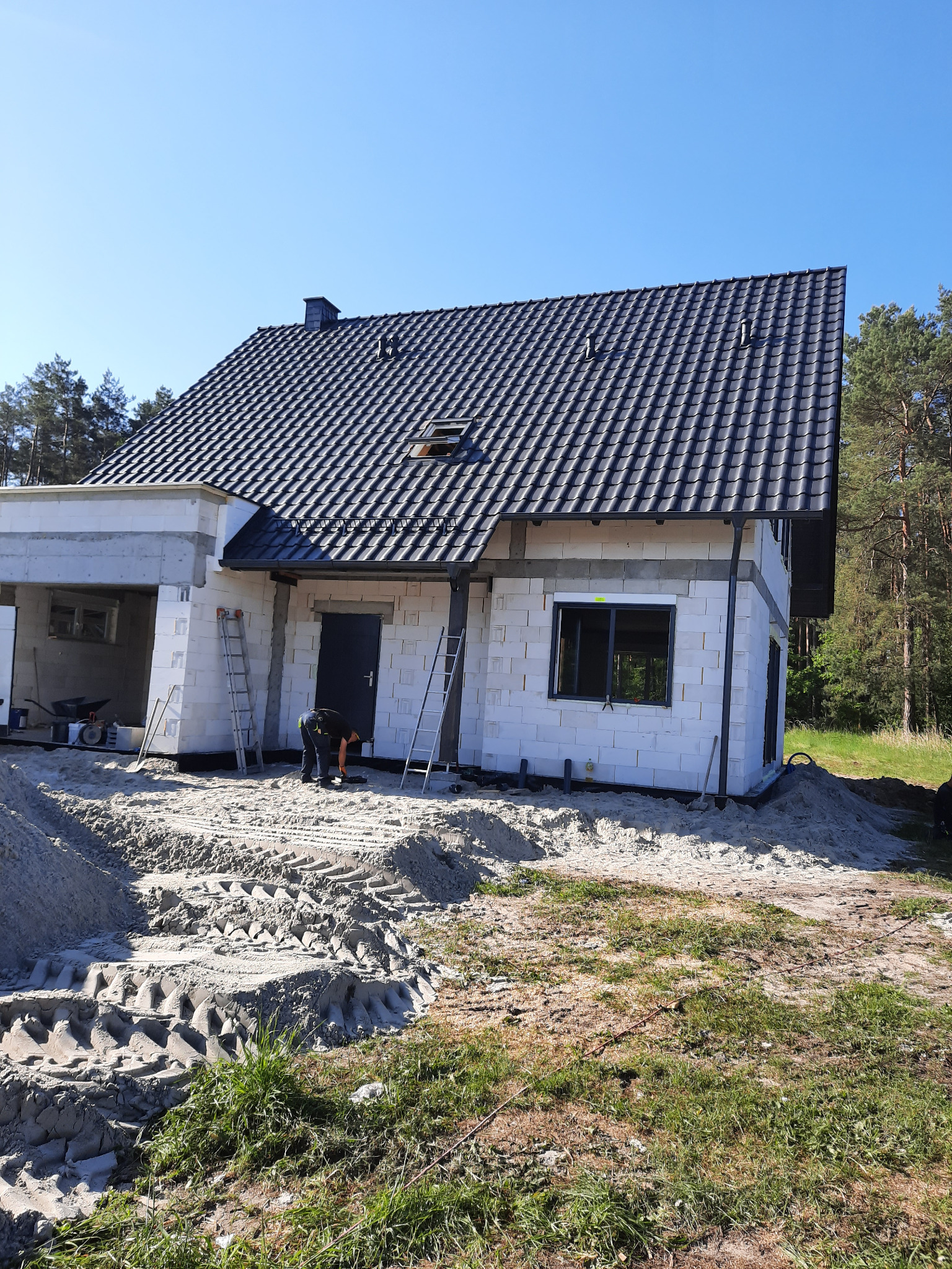 Wznoszony dom z białego bloczka z ciemną dachówką, widoczny pracownik przy wejściu, drabiny oparta o ścianę, teren wokół domu częściowo zasypany piaskiem.