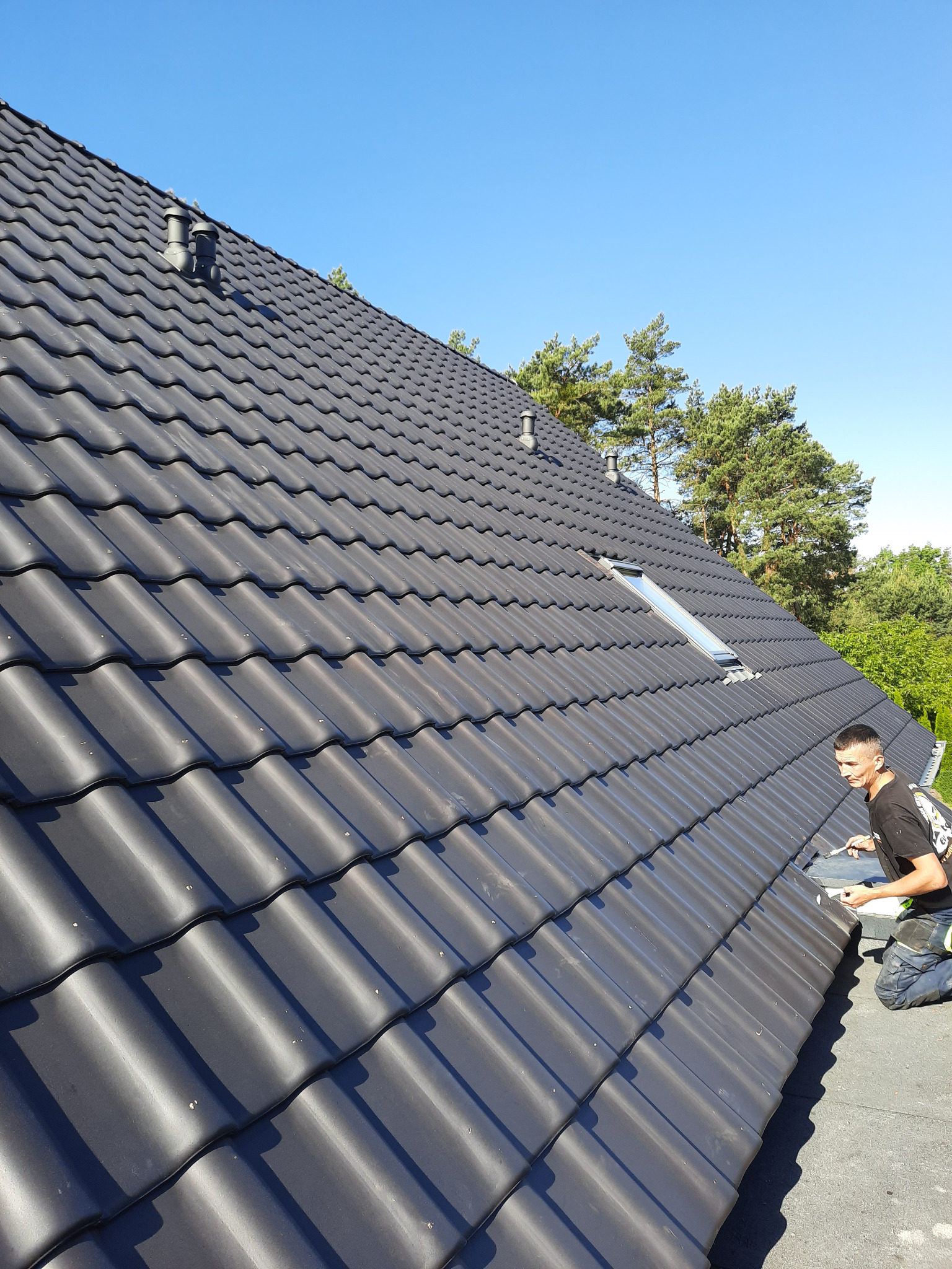 Dekarz na kolanach montuje ciemnoszarą dachówkę na skośnym dachu z oknem dachowym, widoczne kominki wentylacyjne i drzewa w tle pod błękitnym niebem.