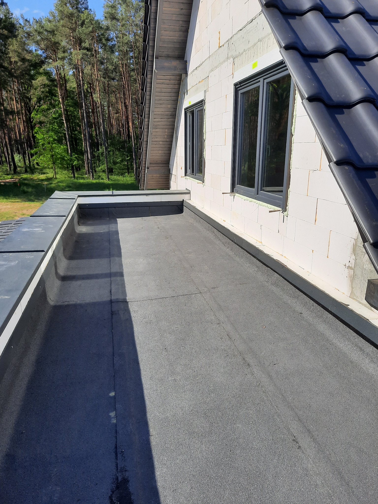 Widok z góry na płaski dach pokryty papą bitumiczną, przylegający do ściany budynku z oknami i fragmentem dachu spadzistego z czarną dachówką, w tle widoczny las.