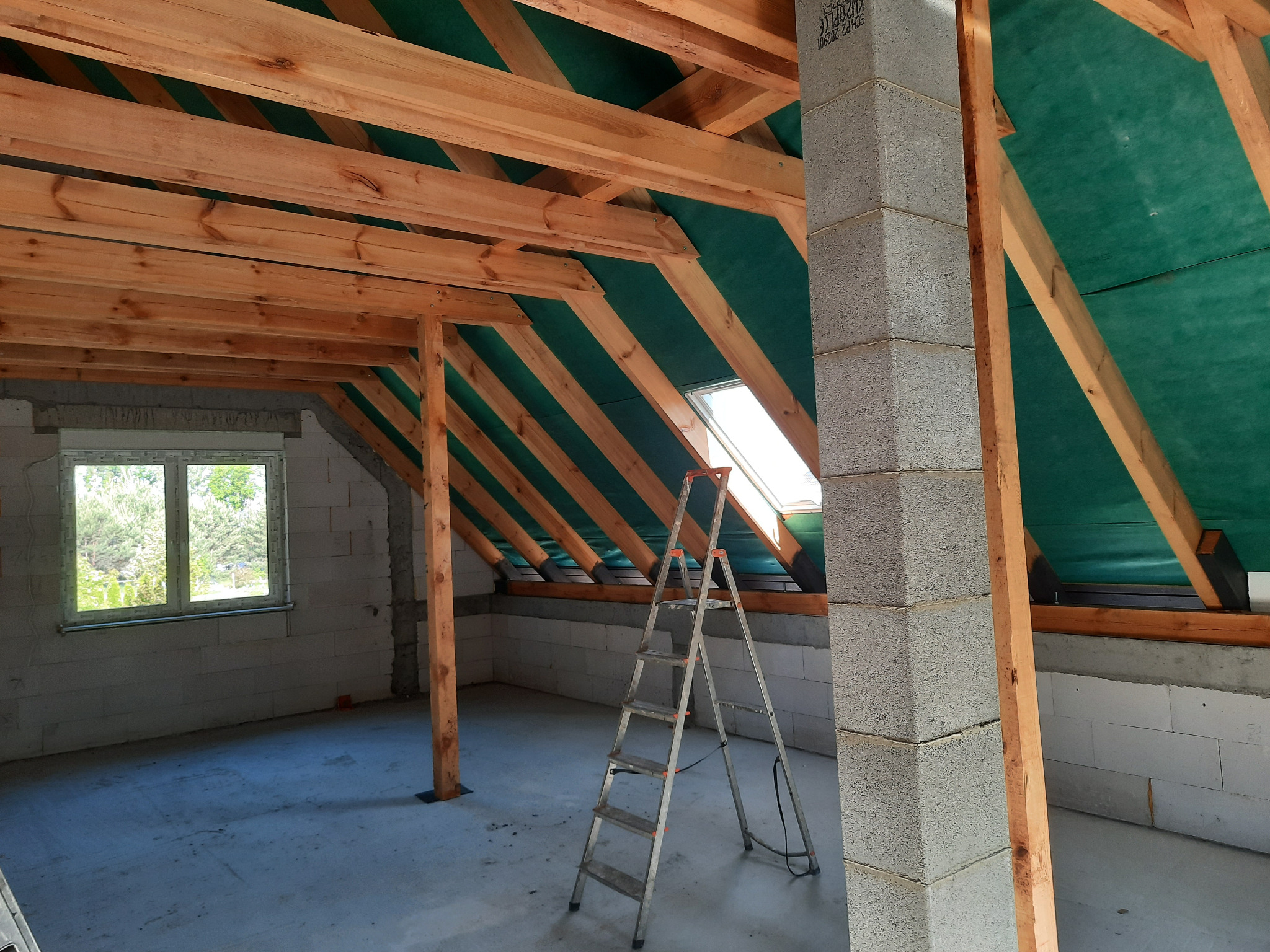 Wnętrze domu w trakcie budowy: widoczne drewniane krokwie dachu pokryte zieloną membraną, ściany z betonu komórkowego, okno i aluminiowa drabina.