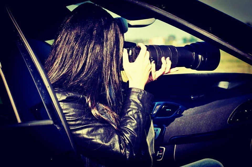 Kobieta w skórzanej kurtce, siedząc w samochodzie, robi zdjęcie profesjonalnym aparatem z dużym teleobiektywem.