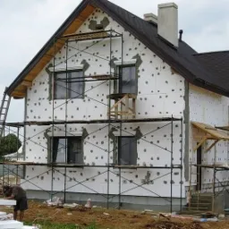 Dom jednorodzinny w trakcie ocieplania styropianem, widoczne rusztowania, pracownicy przygotowujący płyty izolacyjne, otwory po kołkach montażowych, niezabezpieczone elementy drewniane.