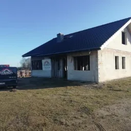 Surowy dom jednorodzinny z ciemnym dachem z dachówki ceramicznej, widoczna elewacja z bloczków betonowych, częściowo zadaszony, na trawiastym terenie, z zaparkowanym vanem firmy dekarskiej...