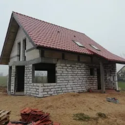 Surowy stan domu jednorodzinnego z czerwoną dachówką, widoczne ściany z bloczków, brak okien i drzwi, palety z dachówką na pierwszym planie, mglista pogoda w tle.