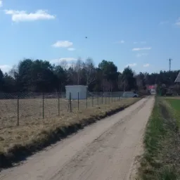 Długa, prosta, polna droga z widokiem na siatkę ogrodzeniową, za którą widać biały budynek i drzewa, w tle domy i wieża telekomunikacyjna.