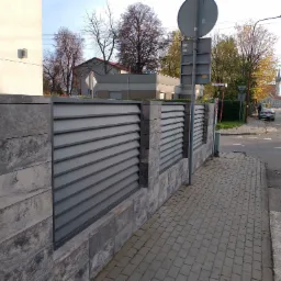 Nowoczesne ogrodzenie panelowe w kolorze szarym z elementami z imitacji kamienia, widok na ulicę i budynki w tle.