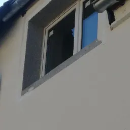 Jasna elewacja domu z oknem obramowanym szarą, kamienną opaską, widoczny fragment rynny.