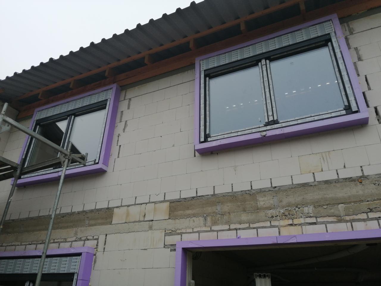Montaż okien z roletami zewnętrznymi w surowym stanie budynku, widoczne rusztowanie i fioletowa izolacja wokół okien.