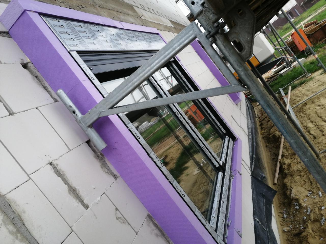 Montaż okna w bloku z białej cegły, ocieplonego fioletowym styropianem, widoczne rusztowanie i teren budowy.