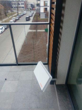 Biała antena satelitarna zamontowana na szarych płytkach balkonu z widokiem na ulicę i budynki mieszkalne w tle.