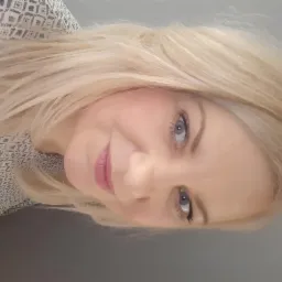 Portret uśmiechniętej kobiety z blond włosami i niebieskimi oczami, ubrana w bluzkę z geometrycznym wzorem.