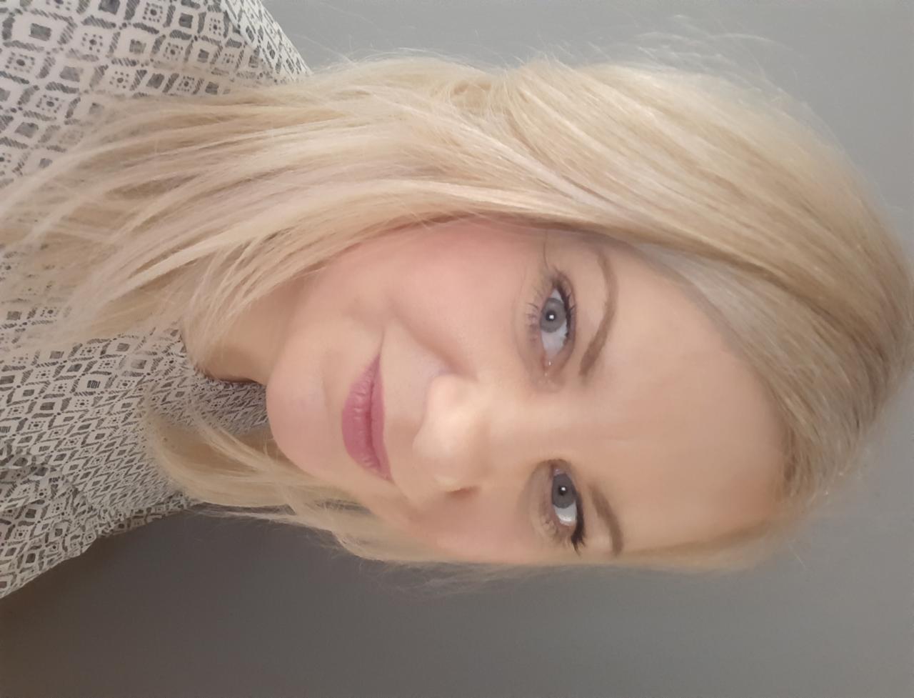 Portret uśmiechniętej kobiety z blond włosami i niebieskimi oczami, ubrana w bluzkę z geometrycznym wzorem.