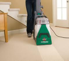 Kobieta w jeansach czyści kremowy dywan za pomocą pionowego, zielono-białego odkurzacza piorącego Bissell. Widoczny fragment schodów i stolika z jasnego drewna w tle.