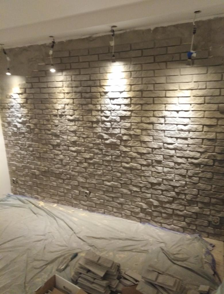 Montaż dekoracyjnej ściany z cegły w pomieszczeniu, widoczne reflektory sufitowe i folia ochronna na podłodze.