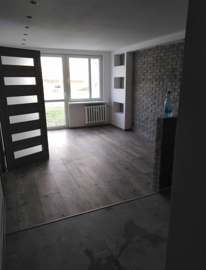 Wykończone wnętrze pokoju z panelami podłogowymi w odcieniach brązu, ścianą z dekoracyjnej cegły, wnękami w ścianie i nowoczesnymi drzwiami z przeszkleniami.