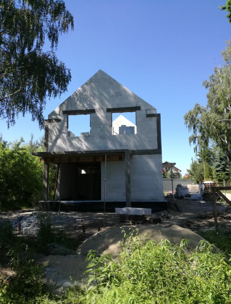 Wznoszony dwukondygnacyjny budynek z białych bloczków, widoczne otwory okienne i zadaszenie wejściowe, na pierwszym planie roślinność i hałda piasku.