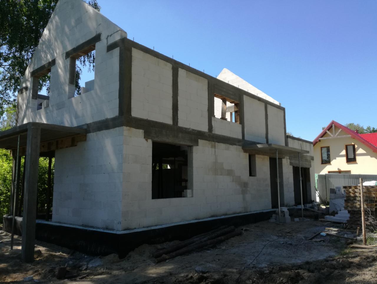 Surowy stan dwukondygnacyjnego domu murowanego z betonu komórkowego, widoczne otwory okienne i drzwiowe, szalunki nadproży, w tle dom jednorodzinny z czerwonym dachem.