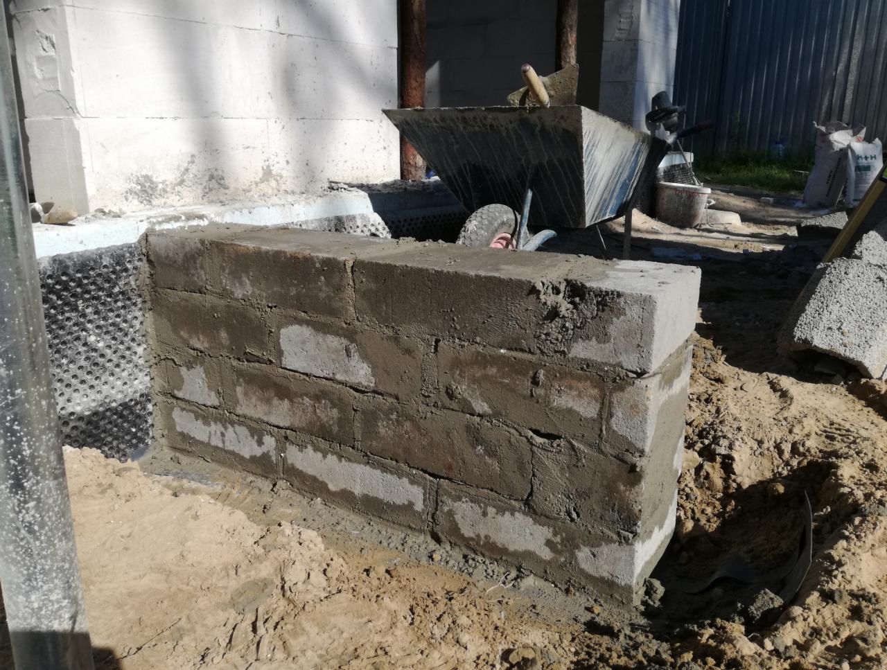Wzniesiony fragment ściany z bloczków betonowych na etapie murowania, widoczna hydroizolacja pionowa fundamentów, taczka z zaprawą w tle.