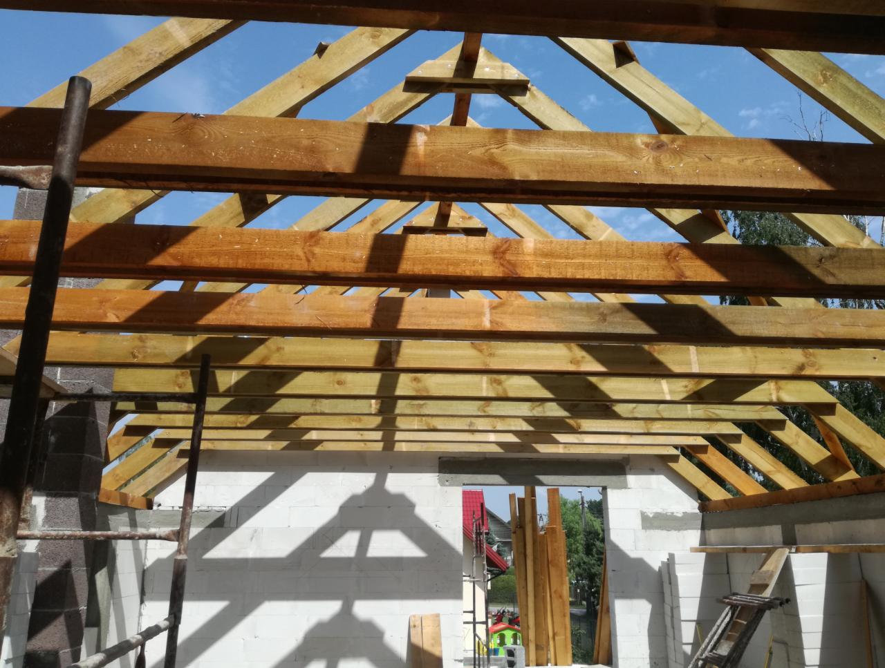 Wnętrze surowego domu z widoczną konstrukcją drewnianą dachu, ściany z betonu komórkowego w trakcie budowy, drabina i rusztowanie w tle.