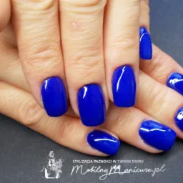 Dłoń z paznokciami pomalowanymi na intensywny, błyszczący, kobaltowy kolor; widoczny logotyp firmy świadczącej usługi mobilnego zdobienia paznokci.