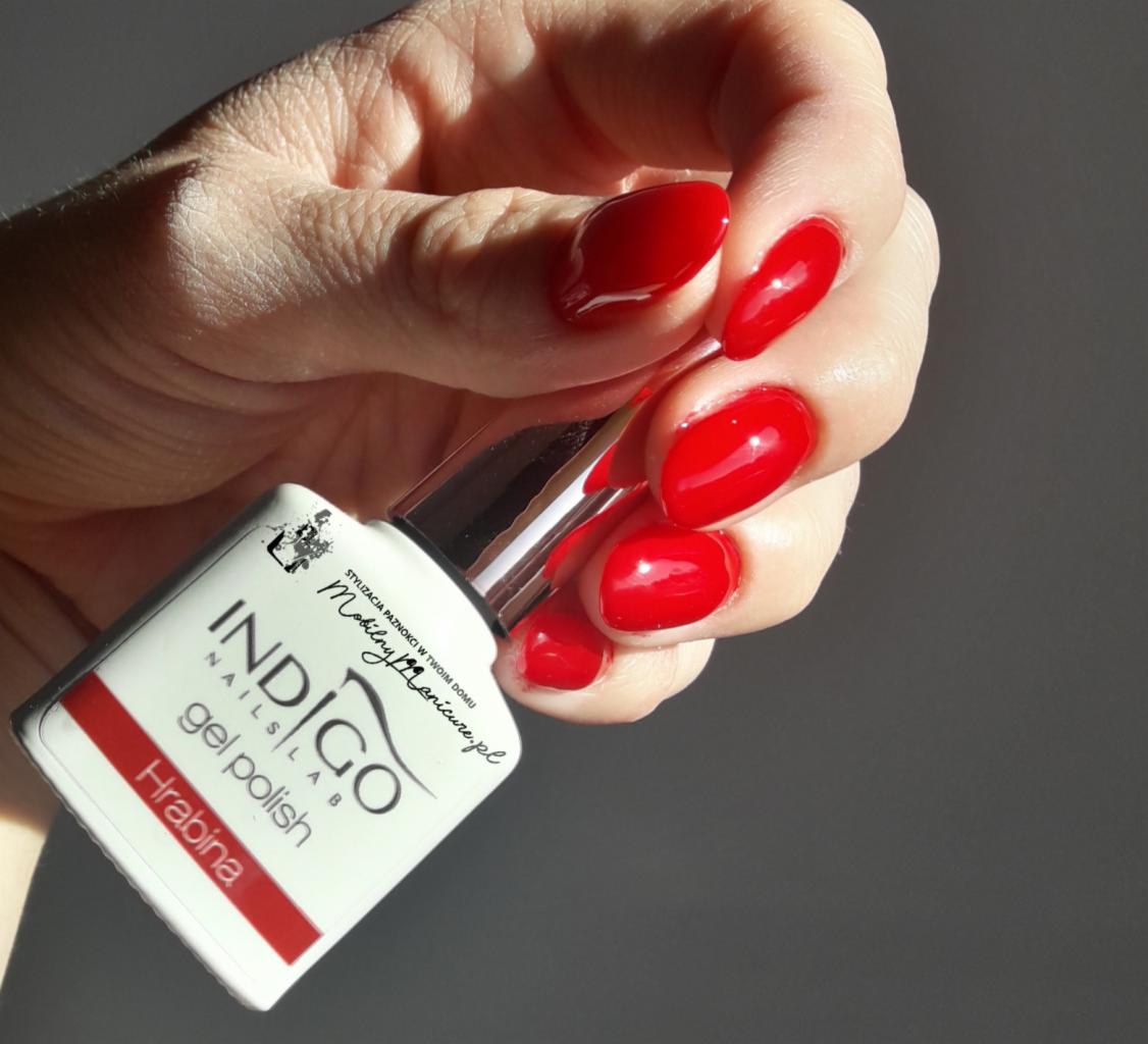 Czerwony lakier hybrydowy Indigo 'Hrabina' na paznokciach dłoni w naturalnym świetle, zbliżenie na buteleczkę i świeżo pomalowane paznokcie.