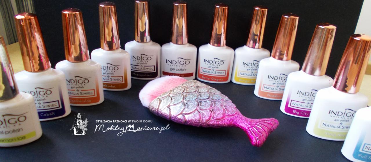 Kolekcja lakierów hybrydowych Indigo w białych buteleczkach z miedzianymi nakrętkami, prezentowana z pędzlem do pyłu w kształcie rybiego ogona na czarnym tle.