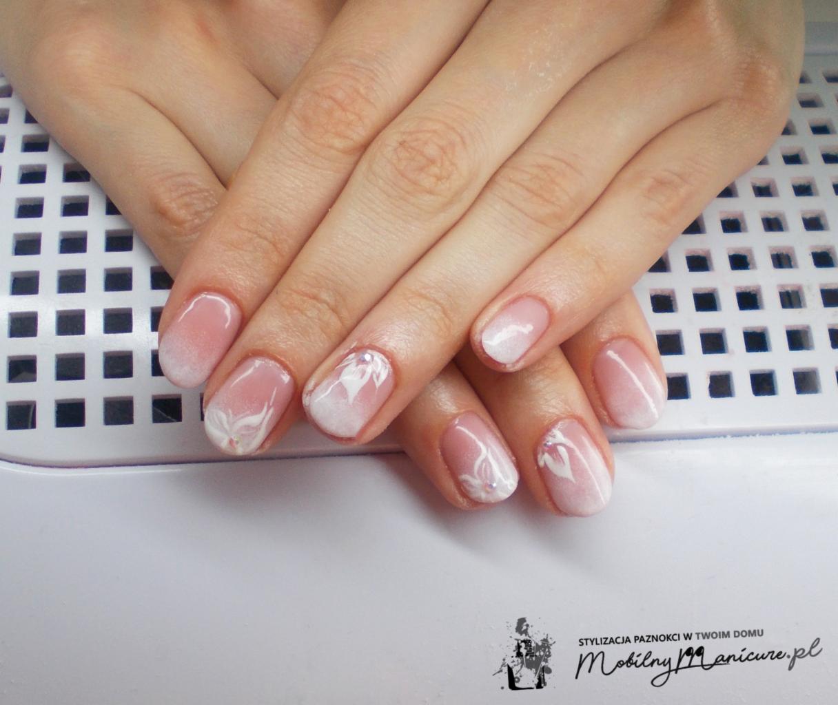 Dłonie z delikatnym, ombre manicure w odcieniach różu i bieli, zdobione białymi kwiatowymi wzorami, ułożone na białej lampie UV.