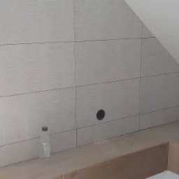 Ściana wyłożona jasnymi, wzorzystymi płytkami ceramicznymi w łazience na poddaszu, z widocznym otworem na gniazdko i butelką wody na drewnianej półce.