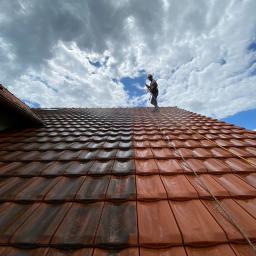 INMAGO Sp. z o.o., Sp. k. - Częściowo umyty dach z czerwonej dachówki, kontrastujące czyste i brudne obszary, pracownik w kasku myje dach pod błękitnym niebem z chmurami.