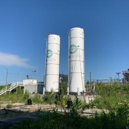 INMAGO Sp. z o.o., Sp. k. - Dwa białe, cylindryczne zbiorniki z logo firmy na tle błękitnego nieba i zabudowań przemysłowych z rurami. W tle widoczne kominy z czerwono-białymi pasami.