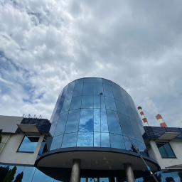 INMAGO Sp. z o.o., Sp. k. - Czyszczenie szklanej elewacji budynku przemysłowego za pomocą długiej lancy teleskopowej. W tle kominy zakładu i pochmurne niebo.