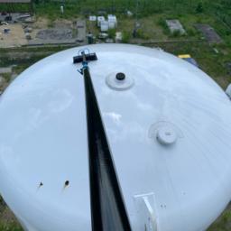 INMAGO Sp. z o.o., Sp. k. - Czyszczenie białego zbiornika przemysłowego z użyciem teleskopowej lancy z szczotką, widoczne ślady zabrudzeń na powierzchni i otoczenie z zielenią w tle.