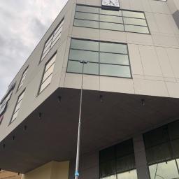 INMAGO Sp. z o.o., Sp. k. - Elewacja budynku biurowego z dużymi oknami, czyszczona za pomocą teleskopowego kija z myjką, widoczny zegar na szczycie budynku i pochmurne niebo w tle.