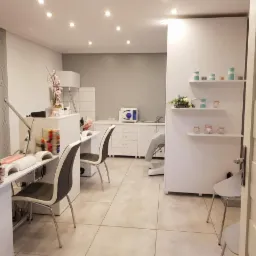 Jasne, przestronne wnętrze gabinetu kosmetycznego z dwoma stanowiskami do manicure, białymi meblami, dekoracyjnymi półkami i sprzętem do sterylizacji.