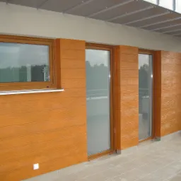 Elewacja budynku z oknem i dwoma przeszklonymi drzwiami balkonowymi, wykończona panelami imitującymi drewno w ciepłym, brązowym odcieniu. Widoczna płytka ceramiczna na balkonie i fragment metalowej...