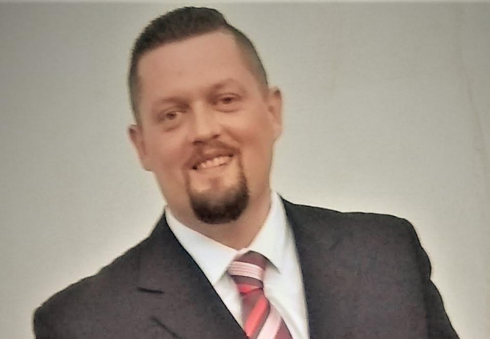 Portret uśmiechniętego mężczyzny w garniturze i krawacie w paski, na jasnym tle.