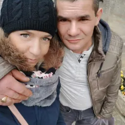 Selfie pary z torbą zakupów na ulicy. Kobieta w czapce i szaliku, mężczyzna w kurtce. Widoczne żółte kwiaty w tle i fragment budynku. Ujęcie z góry.