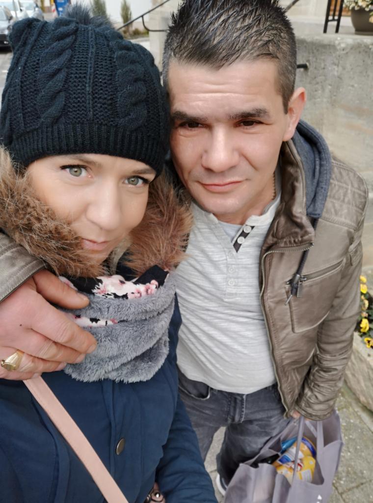 Selfie pary z torbą zakupów na ulicy. Kobieta w czapce i szaliku, mężczyzna w kurtce. Widoczne żółte kwiaty w tle i fragment budynku. Ujęcie z góry.