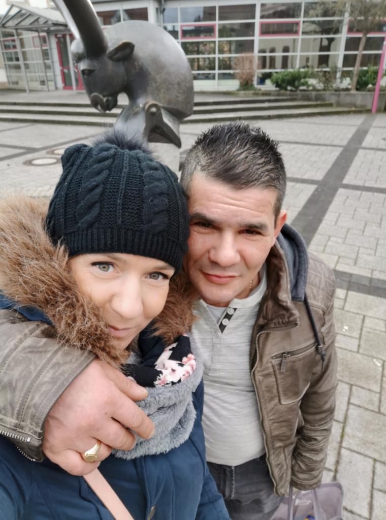 Uśmiechnięta para pozuje do selfie na tle rzeźby nosorożca i budynku z oknami. Mężczyzna w skórzanej kurtce, kobieta w czapce z pomponem i szaliku w kwiaty.