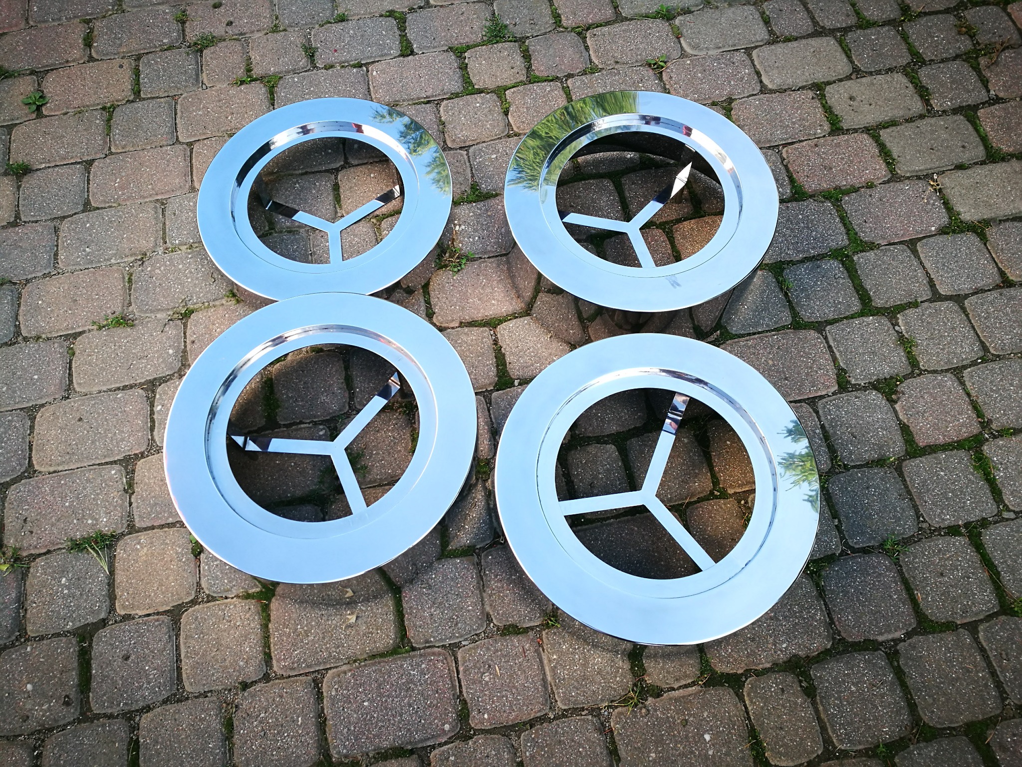 Cztery chromowane, okrągłe elementy konstrukcyjne z trzema wspornikami wewnątrz, leżące na brukowanej powierzchni.