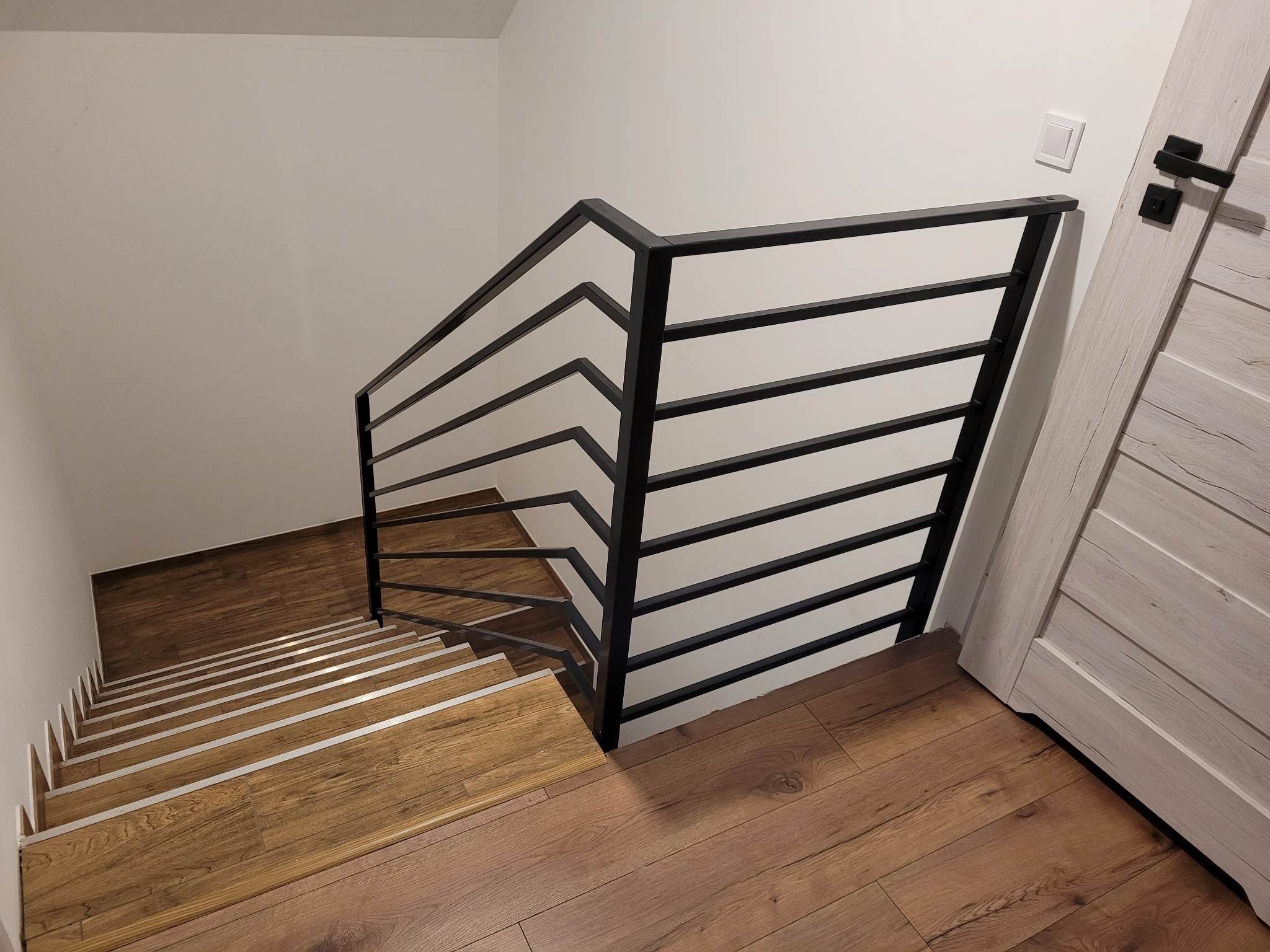 Nowoczesne schody z drewnianymi stopniami i czarną, poziomą balustradą z metalowych prętów, widoczne z góry, z fragmentem drewnianej podłogi i drzwi w tle.