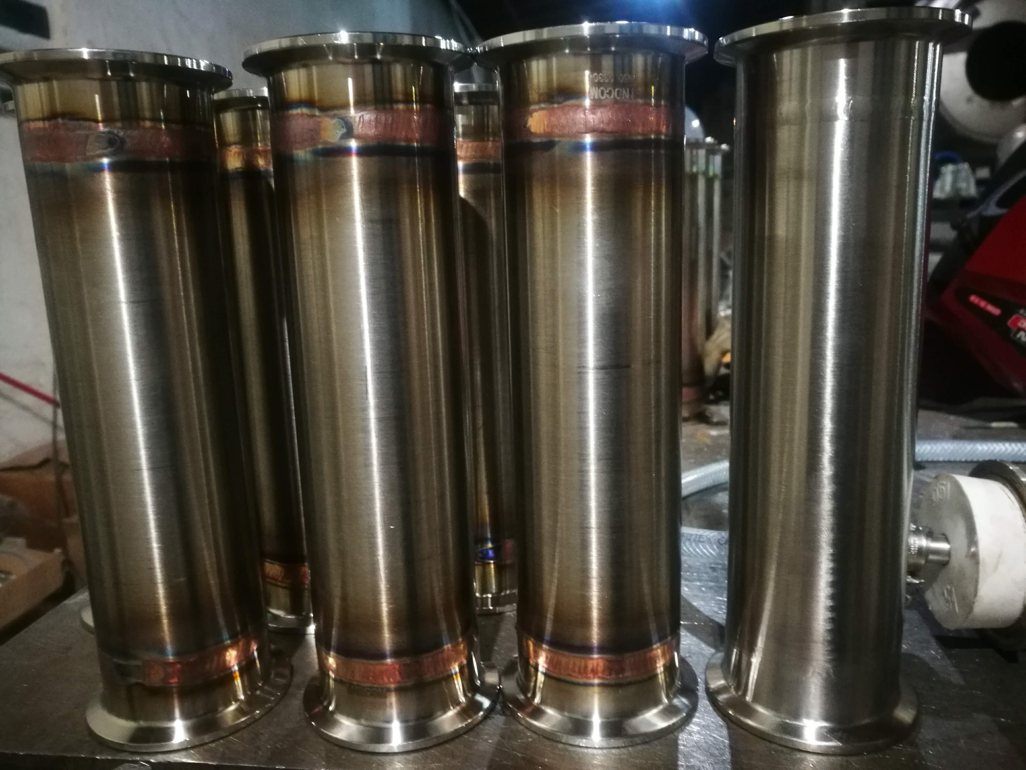 Szereg polerowanych, metalowych rur z widocznymi śladami łączenia, z opalizującymi przebarwieniami po obróbce termicznej, ustawionych na metalowym stole.