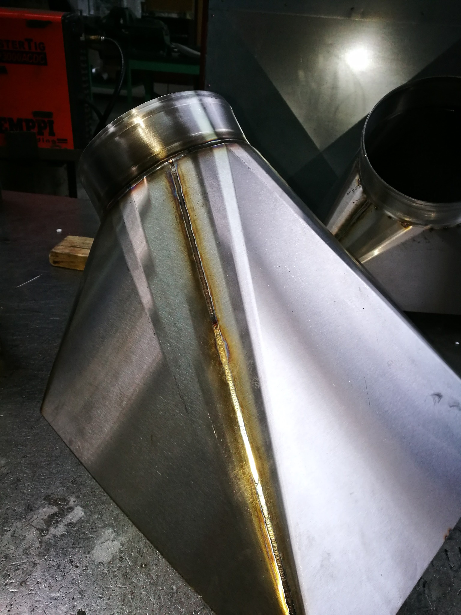 Spoina na elemencie ze stali nierdzewnej, widoczna tęczowa barwa spoiny, w tle sprzęt spawalniczy.