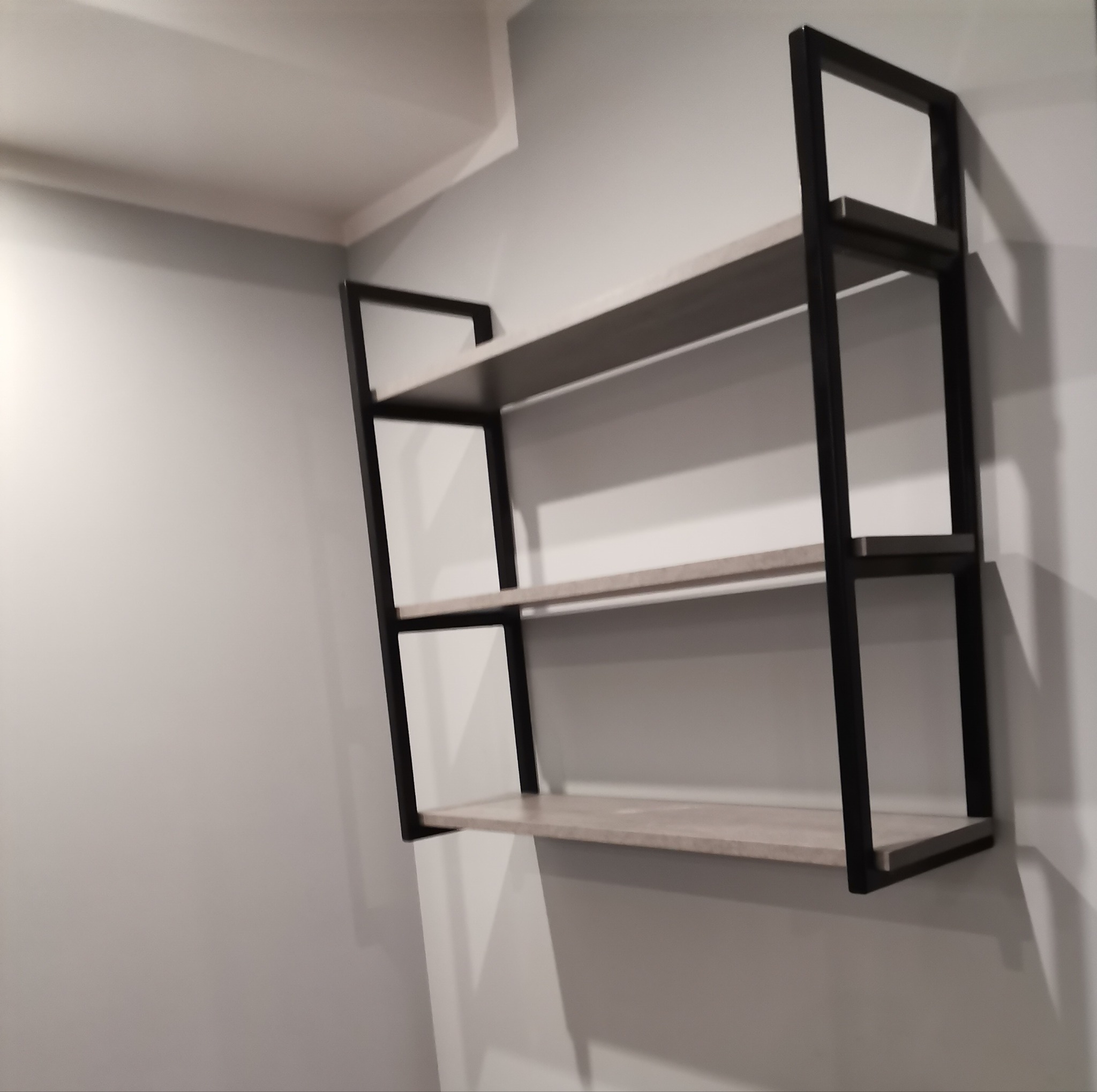 Trzy półki z czarną metalową ramą przymocowane do szarej ściany, widoczne z boku.