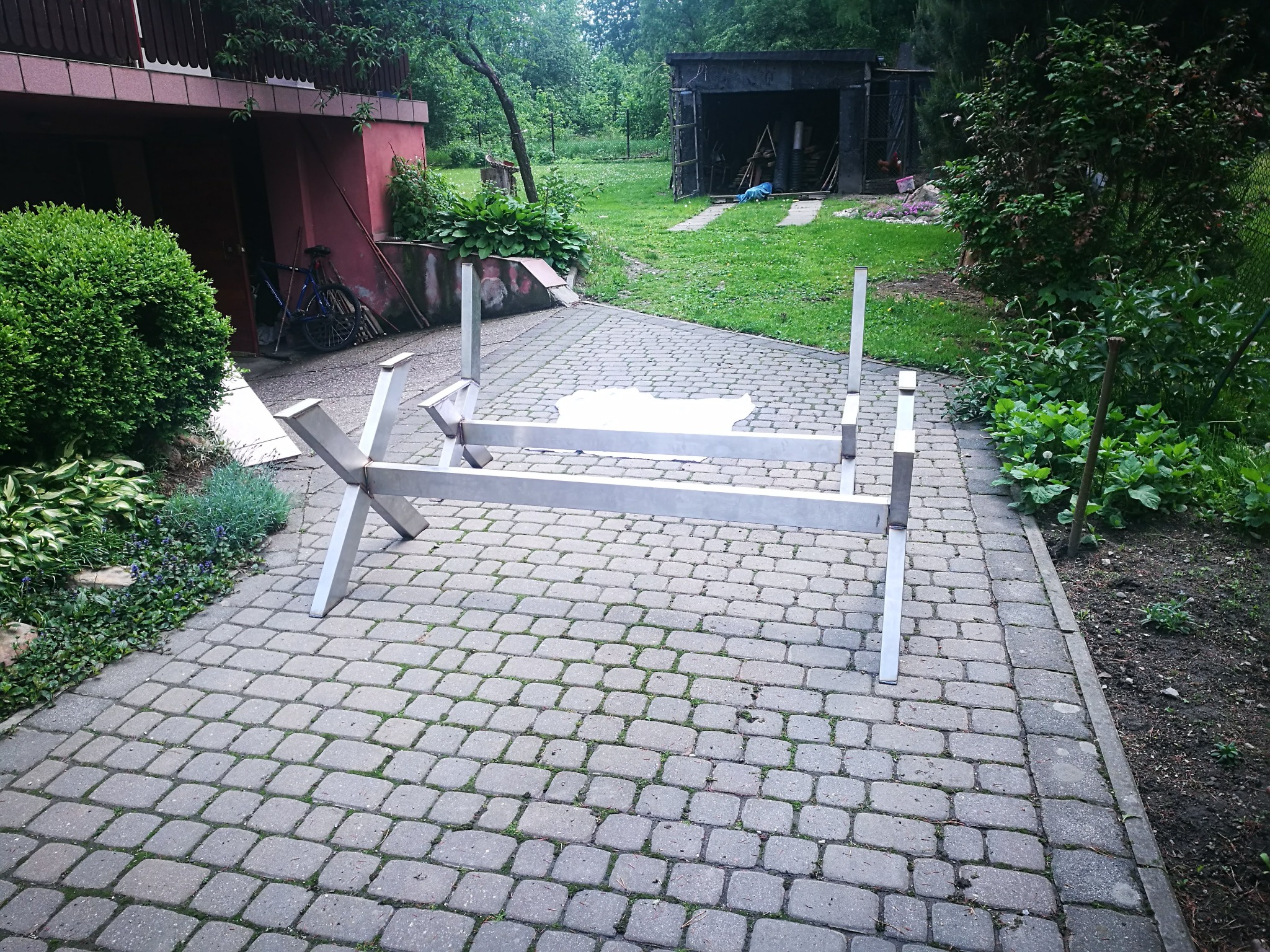 Metalowa konstrukcja spawana z profili, prawdopodobnie podstawa stołu, leży na brukowanej nawierzchni w ogrodzie.