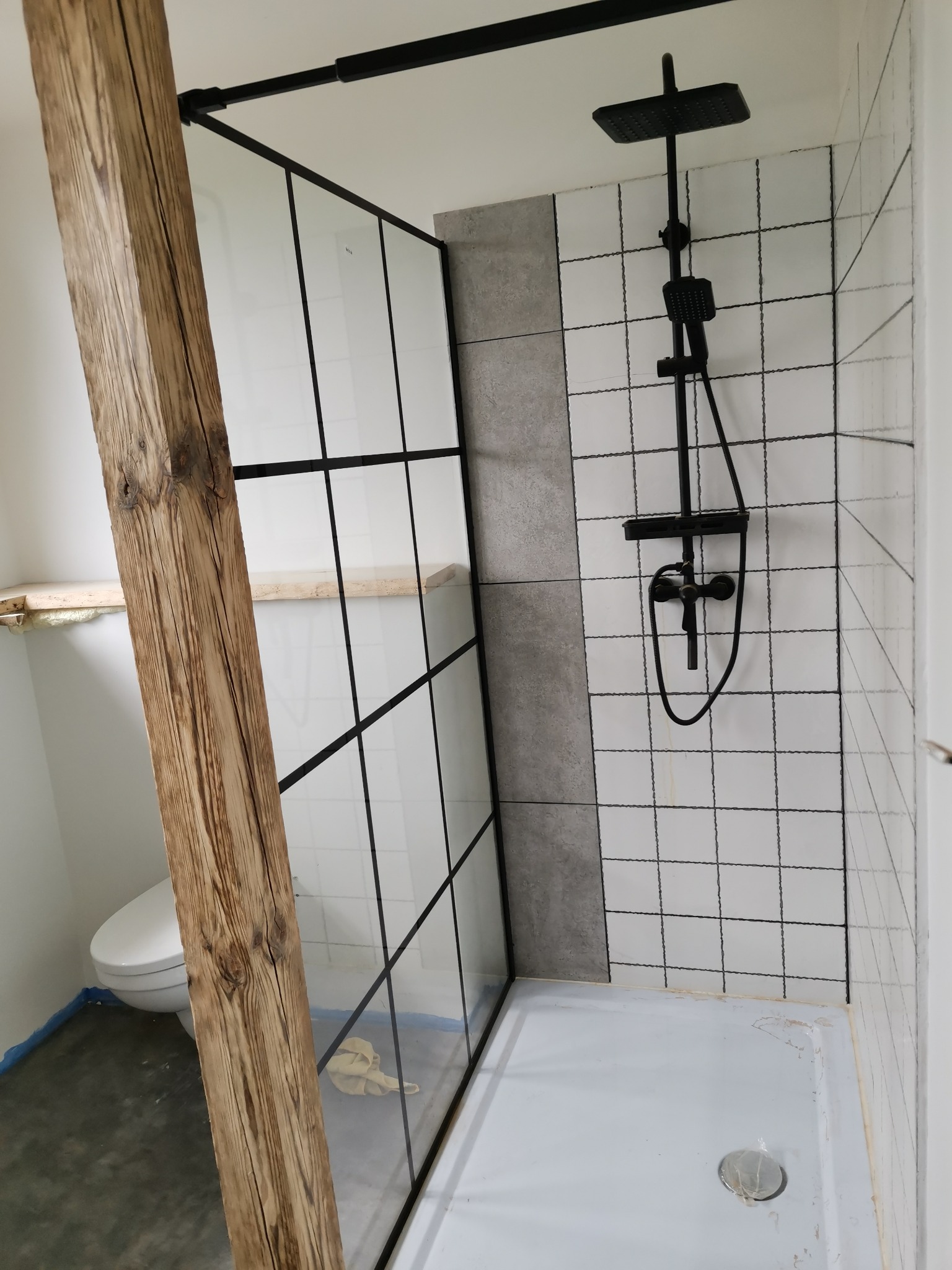 Kabina prysznicowa z czarnymi profilami i drewnianą belką, białe płytki z czarną fugą, czarna armatura, widoczny fragment toalety, podłoga zabezpieczona folią.