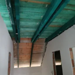 Poddasze w trakcie prac wykończeniowych: widoczne drewniane belki pomalowane na zielono oraz surowe ściany z widocznymi fragmentami cegieł wokół otworów drzwiowych.